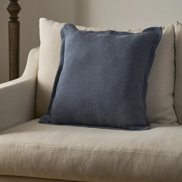 A square blue throw pillow with a linen-like texture, styled on a beige upholstered armchair.