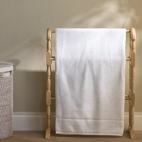 A white bath towel with a decorative border hangs from a light natural wood towel rack, set against a textured beige wall with a woven basket partially visible on the left.