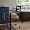A folded stack of soft white bath sheets,  placed on the woven seat of a dark wooden chair in a warm indoor setting with wood flooring and a rug, in a traditional bathroom.