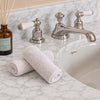 Two rolled white flannels placed on a marble bathroom counter beside a chrome faucet and sink, with a glass reed diffuser visible at left.
