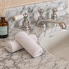 Two rolled white flannels placed on a marble bathroom counter beside a chrome faucet and sink, with a glass reed diffuser visible at left.