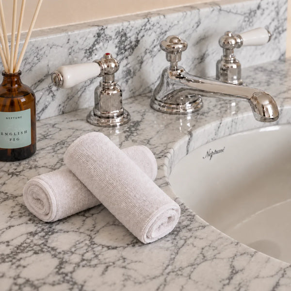 Two rolled white flannels placed on a marble bathroom counter beside a chrome faucet and sink, with a glass reed diffuser visible at left.