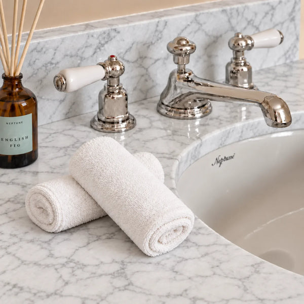 Two rolled white towels on a white marble bathroom counter beside a polished chrome faucet and a small amber reed diffuser bottle.