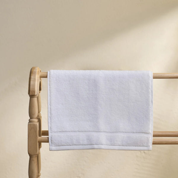 A white cotton hand towel with a stitched border hangs from a light natural wood towel rack against a warm beige wall.