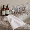A folded white cotton guest towel with a stitched border rests on a marble bathroom countertop beside two amber pump bottles labeled English Fig and a polished chrome faucet.