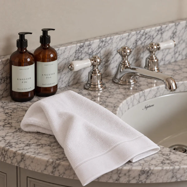 A folded white cotton guest towel with a stitched border rests on a marble bathroom countertop beside two amber pump bottles labeled English Fig and a polished chrome faucet.