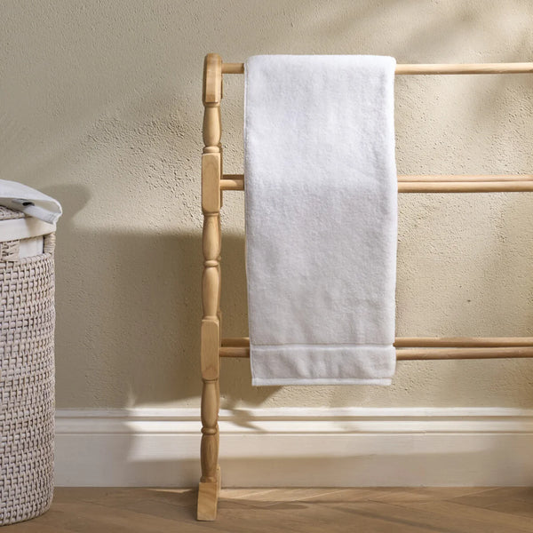 A white cotton hand towel with a folded hem is draped over a light natural wood towel rack with turned spindles, set against a textured beige wall and light wood floor.