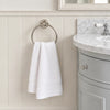 A white hand towel hangs from a round silver towel ring mounted on a cream paneled bathroom wall, with part of a gray vanity and marble countertop visible on the right.