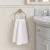 A white hand towel hangs from a round silver towel ring mounted on a cream paneled bathroom wall, with part of a gray vanity and marble countertop visible on the right.