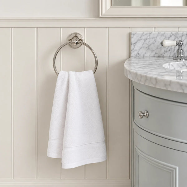 A white hand towel hangs from a round silver towel ring mounted on a cream paneled bathroom wall, with part of a gray vanity and marble countertop visible on the right.