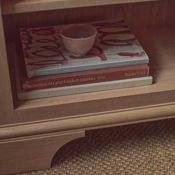 A close-up of a wooden shelf holding a stack of books and a small pink bowl. Visible book titles include 'Decorative Art and Modern Interiors 1980' and 'Studio Vista'.