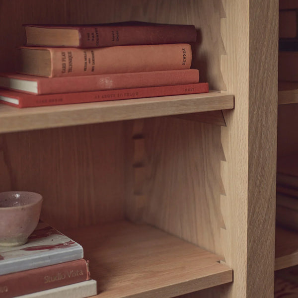 Close-up of a light natural wood bookshelf with stacked books on the shelves, including a small ceramic bowl on the lower shelf.