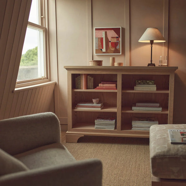 A warm, styled interior scene featuring a light wood bookcase with books and decor, a table lamp, wall art, a window, and parts of upholstered furniture in a cozy room setting.
