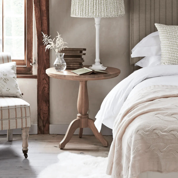 A round wooden nightstand in a rustic bedroom setting, styled with a white lamp, a small glass vase with dried stems, and a stack of books, positioned next to a bed with white linens.
