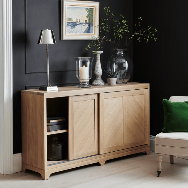 A light oak wood sideboard cabinet with two central doors and an open shelf on the left, styled in a modern living room setting against a dark wall with decorative vases, a lamp, and framed art.