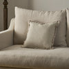 A square beige throw pillow with a fringed edge resting on a matching upholstered armchair in a softly lit interior setting.