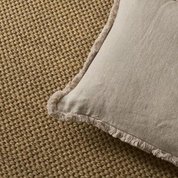 A close-up of a light beige linen throw pillow with fringed edges, resting on a textured brown woven rug.