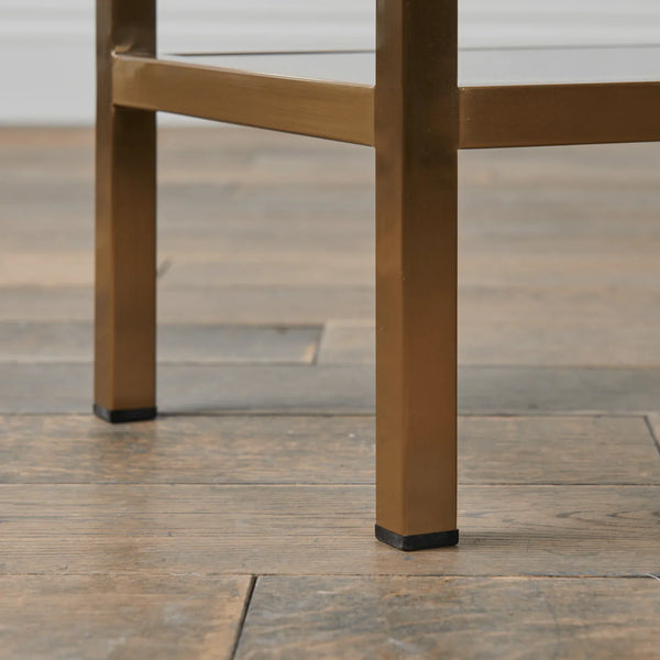 Close-up of a brass-finished metal table leg with a black rubber foot cap, standing on a rustic wooden plank floor.
