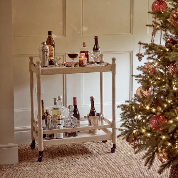 A two-tier wooden bar cart with wheels, styled with various liquor bottles, glasses, and a candle, set in a decorated room next to a Christmas tree.