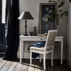 A styled interior scene showing a white writing desk with a matching upholstered chair, a black table lamp, a vase, books, and framed wall art in a room with dark curtains and wood flooring.