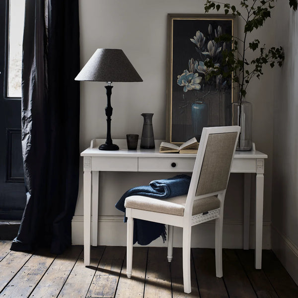 A styled interior scene showing a white writing desk with a matching upholstered chair, a black table lamp, a vase, books, and framed wall art in a room with dark curtains and wood flooring.