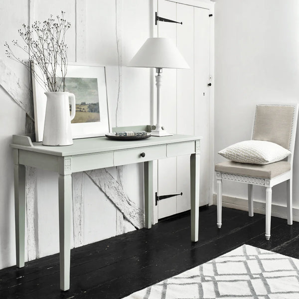 A pale sage green writing desk with a single drawer and decorative legs, styled in a bright, modern room next to a beige upholstered chair. The desk is decorated with a white pitcher of dried stems, a framed print, and a white table lamp.