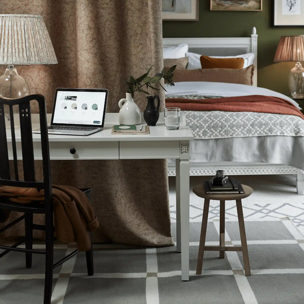 A styled bedroom interior featuring a white writing desk with a laptop, vase, and glass, positioned in front of a brown curtain. A black wooden chair sits on the left, and a small round wooden stool is on the right.