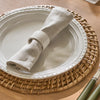 A rolled beige linen napkin with a white fabric napkin ring, set on a white ceramic plate with a woven round placemat on a wooden table.