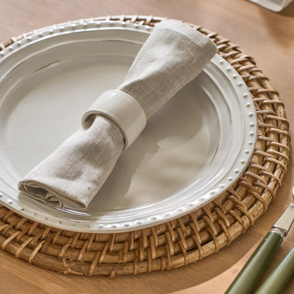 A rolled beige linen napkin with a white fabric napkin ring, set on a white ceramic plate with a woven round placemat on a wooden table.