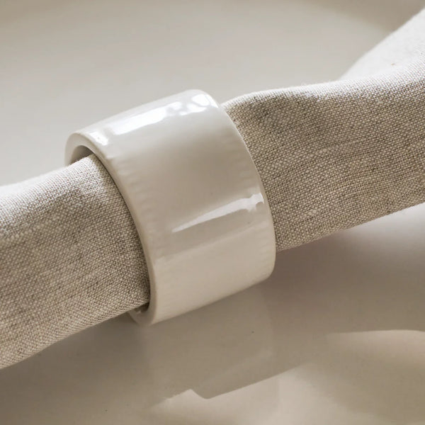 A glossy white ceramic napkin ring holding a textured beige linen napkin, shown in a close-up studio shot.