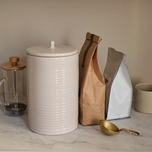 A ribbed ceramic canister with a matching lid sits on a marble countertop next to a glass French press, a brown paper bag, a white bag, and a gold scoop, against a neutral beige wall.