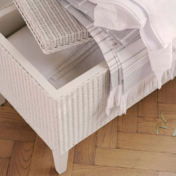 A white woven rattan storage bench with an open drawer, styled with a white waffle-knit throw blanket and striped pillow on a light wood parquet floor.