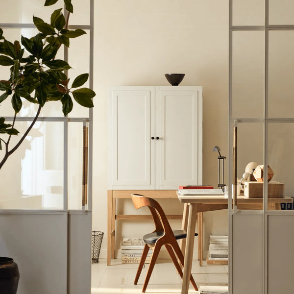A minimalist interior scene featuring a white double-door cabinet, a light wood desk, a wooden chair, and a potted plant, framed by a glass-paneled screen in a bright, modern room.