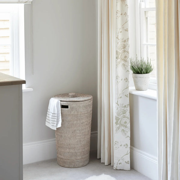 A tall woven natural-fiber laundry hamper with a lid stands in a bright room beside a window with cream floral curtains. A white towel with thin black stripes hangs from the side, and a small potted plant sits on the windowsill.