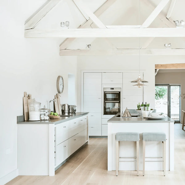 A bright, modern kitchen interior with white cabinetry, a white island, gray stone countertops, light wood flooring, and two light blue upholstered bar stools at the island.