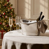 A silver metal ice bucket filled with ice and two wine bottles, set on a white tablecloth next to two champagne flutes. A decorated Christmas tree with warm lights is blurred in the background.