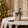 Silver wine cooler filled with ice and a champagne bottle, accompanied by two champagne flutes and folded linen napkins on a table, with a softly lit, decorated Christmas tree in the background.