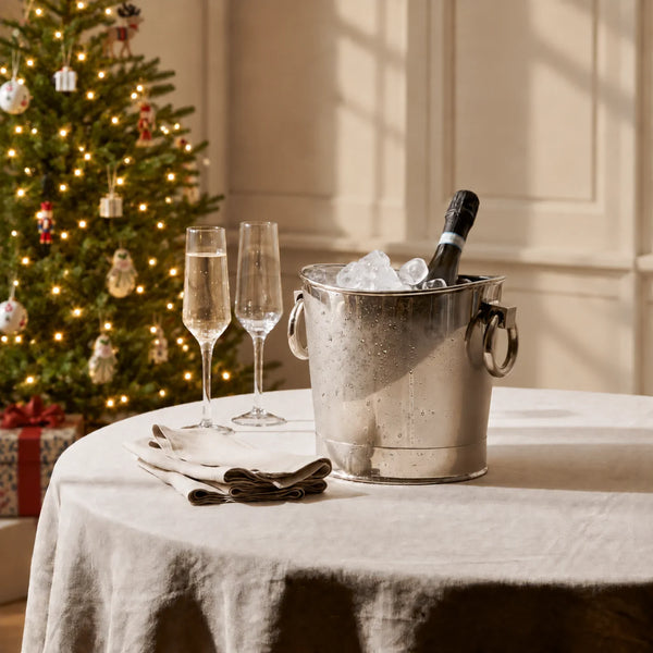 Silver wine cooler filled with ice and a champagne bottle, accompanied by two champagne flutes and folded linen napkins on a table, with a softly lit, decorated Christmas tree in the background.