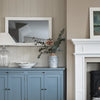 A styled interior scene showing a blue cabinet against a beige paneled wall, with a white-framed mirror, a glass lamp, a bowl, and a vase with greenery. Part of a white fireplace and a framed landscape print are visible on the right.