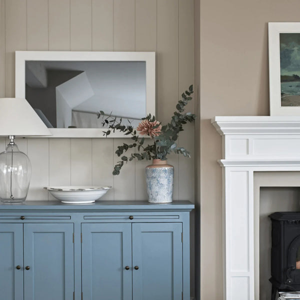 A styled interior scene showing a blue cabinet against a beige paneled wall, with a white-framed mirror, a glass lamp, a bowl, and a vase with greenery. Part of a white fireplace and a framed landscape print are visible on the right.