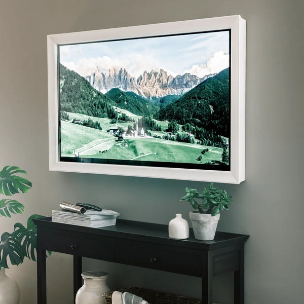 A white wall-mounted flat-screen TV with a black bezel displaying a mountain landscape, mounted on a gray wall above a black console table with decorative plants and vases.