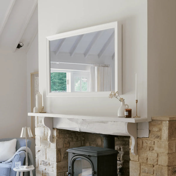 A large white-framed rectangular mirror hangs on a white wall above a stone fireplace mantel in a bright, modern living room.