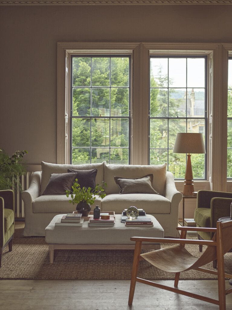 A styled living room featuring a cream-colored fabric sofa with dark brown accent pillows, set in front of a large multi-pane window. A matching cream coffee table sits in the foreground, alongside a wooden chair, a floor lamp, and a woven rug, with lush green trees visible outside the windows.