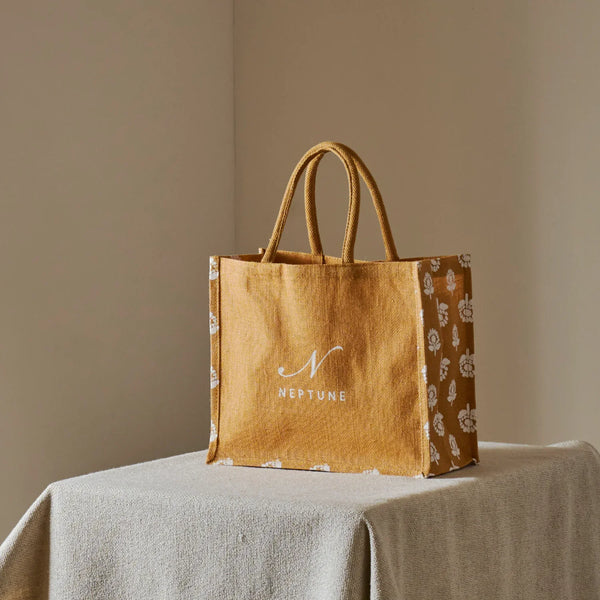 A mustard yellow canvas tote bag with white floral patterned sides and a white printed 'N' logo and 'NEPTUNE' text on the front, displayed on a draped light beige tablecloth against a neutral beige background.