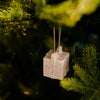 A small speckled off-white ceramic Christmas ornament shaped like a miniature house hangs from a pine tree branch.