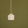 A small gift box pendant hanging from a thin cord against a solid olive green background. The pendant is off-white with brown speckles and a matching bow detail.