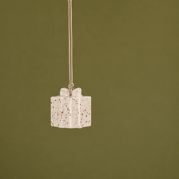 A small gift box pendant hanging from a thin cord against a solid olive green background. The pendant is off-white with brown speckles and a matching bow detail.