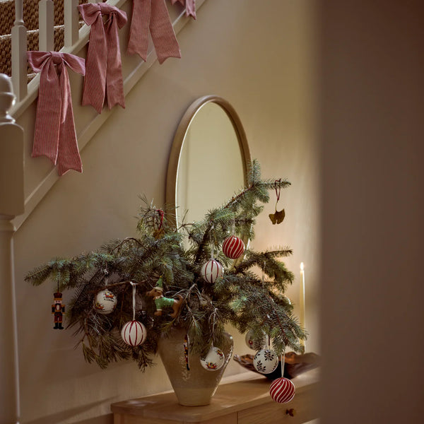 A small decorative Christmas tree in a beige vase on a wooden console table, decorated with red-and-white striped ornaments and hanging ornaments, set in a warmly lit entryway with a mirror and pink stair rail bows in the background.