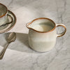 Cream ceramic milk jug with brown glazed rim on a marble tabletop beside a spoon and cup.