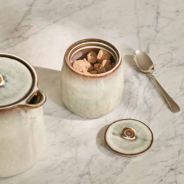 Cream ceramic sugar pot with brown glazed rim, filled with brown sugar cubes on a marble tabletop.